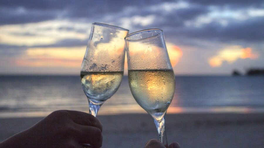 Two glasses of white wine clinking together, held by two hands, with a scenic beach and ocean backdrop during a colorful sunset.