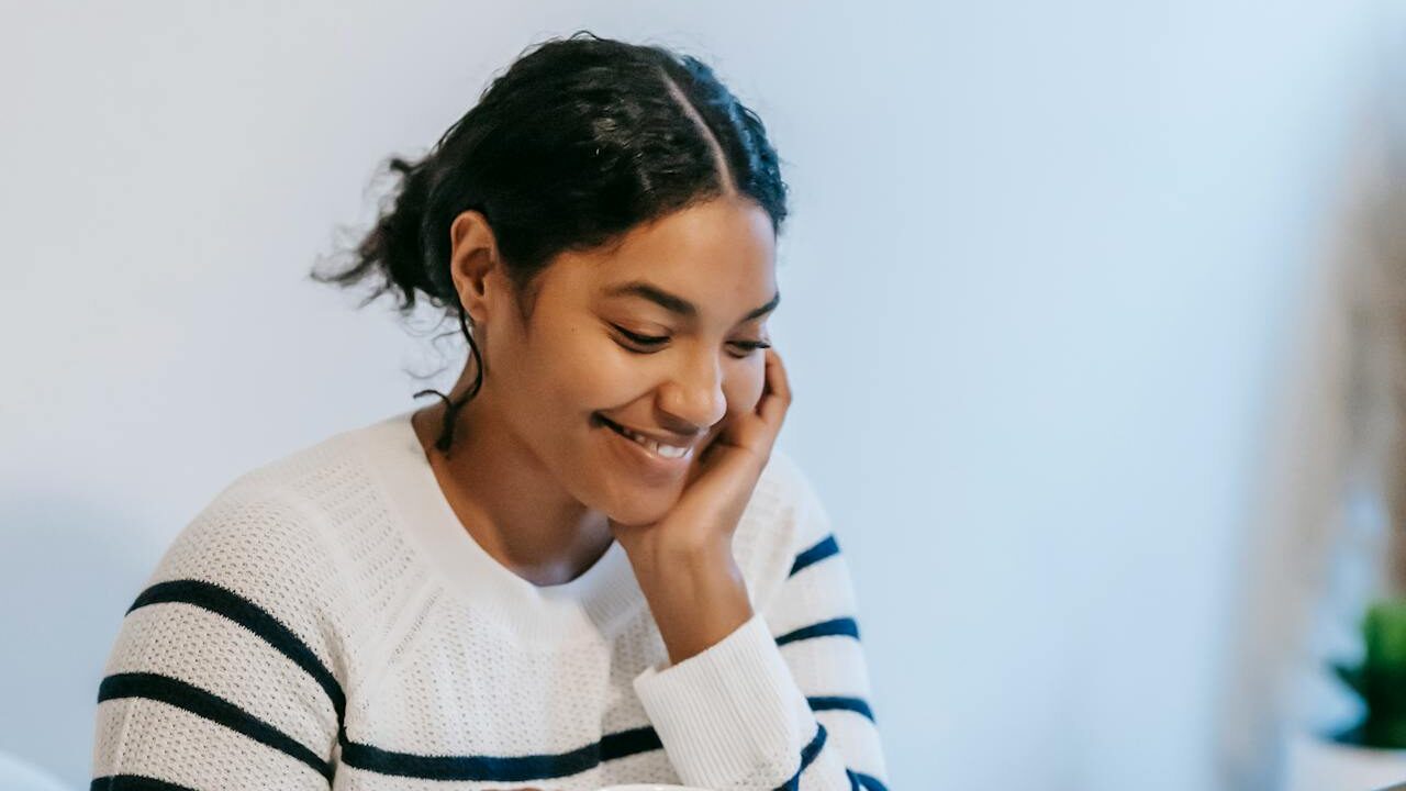 A smiling woman with curly dark hair, wearing a white sweater with navy blue stripes, resting her head on her hand in a softly lit indoor setting with a light blue background.