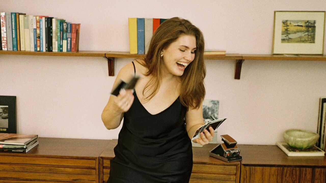 A cheerful woman dressed in a black slip dress, holding polaroid photos, standing in front of a wall-mounted bookshelf and vintage-style furniture in a warmly lit space.