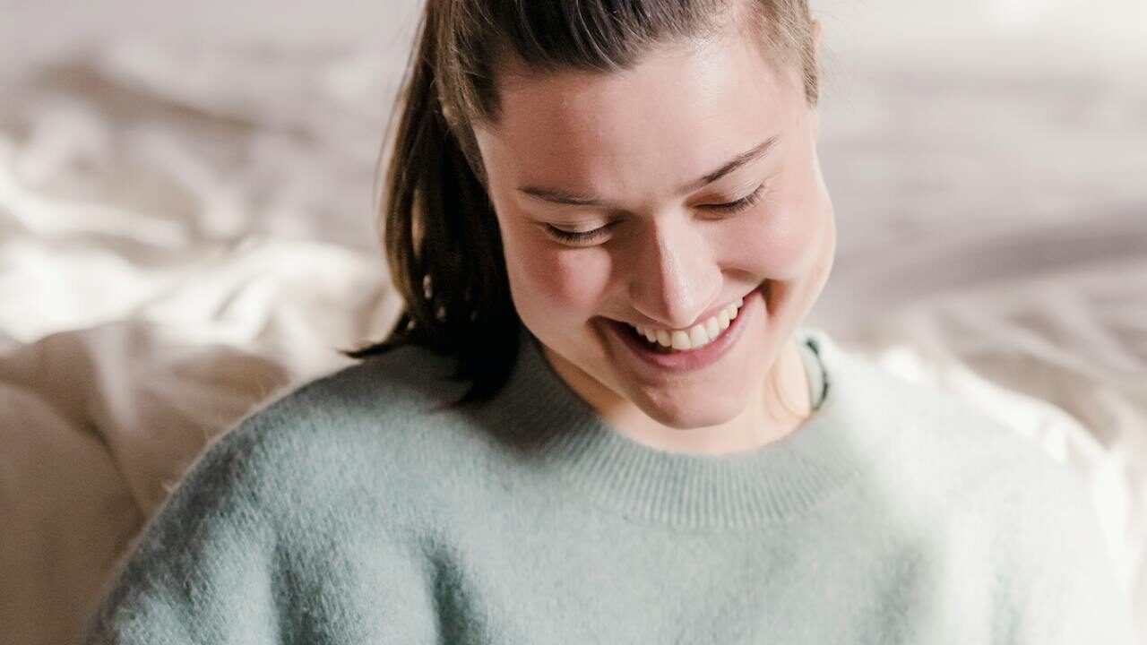 A cheerful woman with light skin and tied-back hair, captured in a candid moment of laughter, wearing a soft green sweater in a relaxed environment.