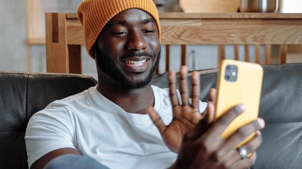 Man wearing a beanie and white shirt waving at his phone during a video call, seated on a couch.