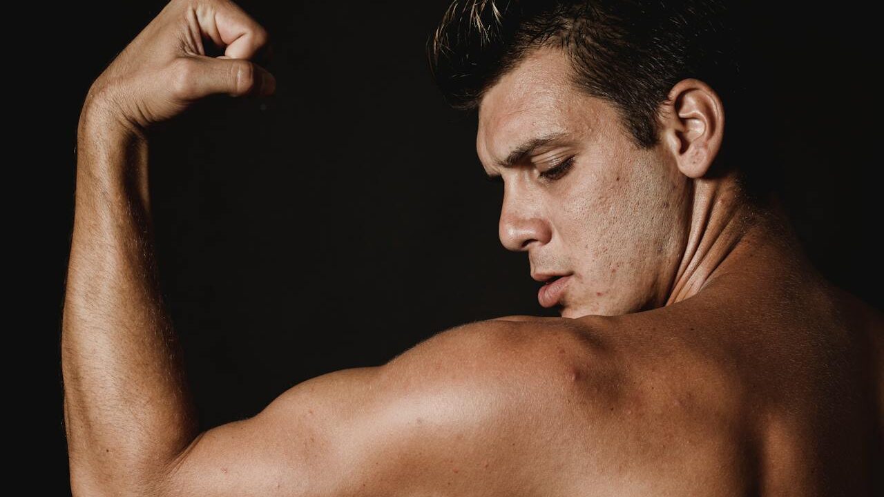 Shirtless man flexing his bicep, looking at his arm against a dark background.