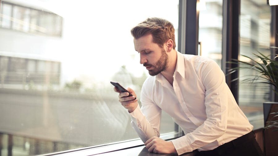 A man in a white shirt leaning by a window, looking at his smartphone.