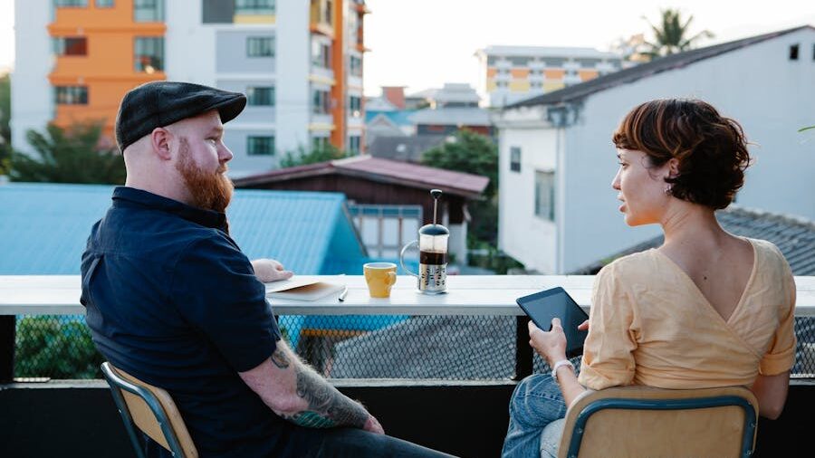 Two people sitting on a balcony table, engaged in conversation with coffee and a tablet.