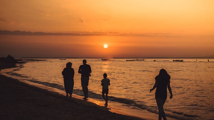 Four people strolling along a sandy beach, with boats in the distance and a glowing orange sunset.