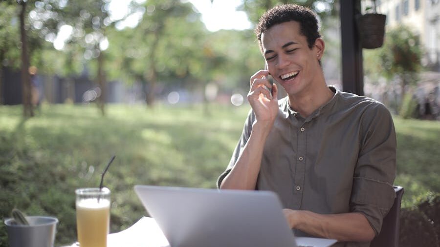 Smiling person sitting at an outdoor table, talking on the phone and using a laptop.