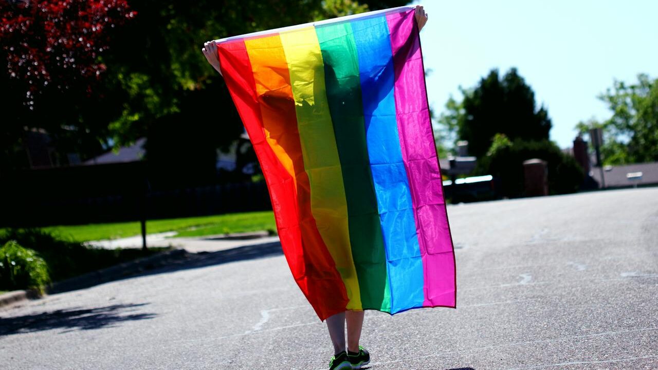 A person holding a rainbow flag behind them while walking on a sunny street.