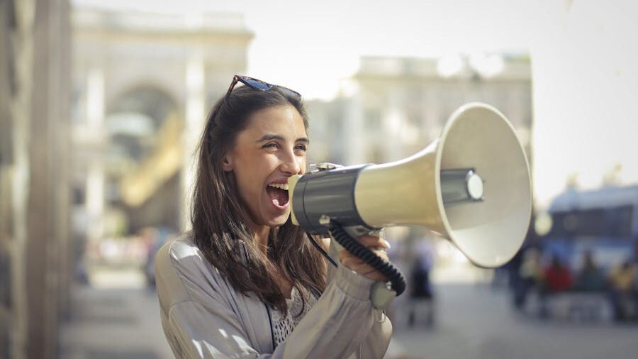 Woman using a megaphone in a city setting with people and buildings in the background.