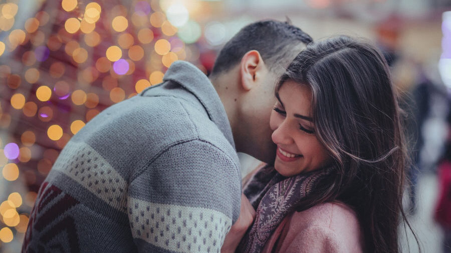 A man embracing a smiling woman in front of a decorated Christmas tree with glowing lights.