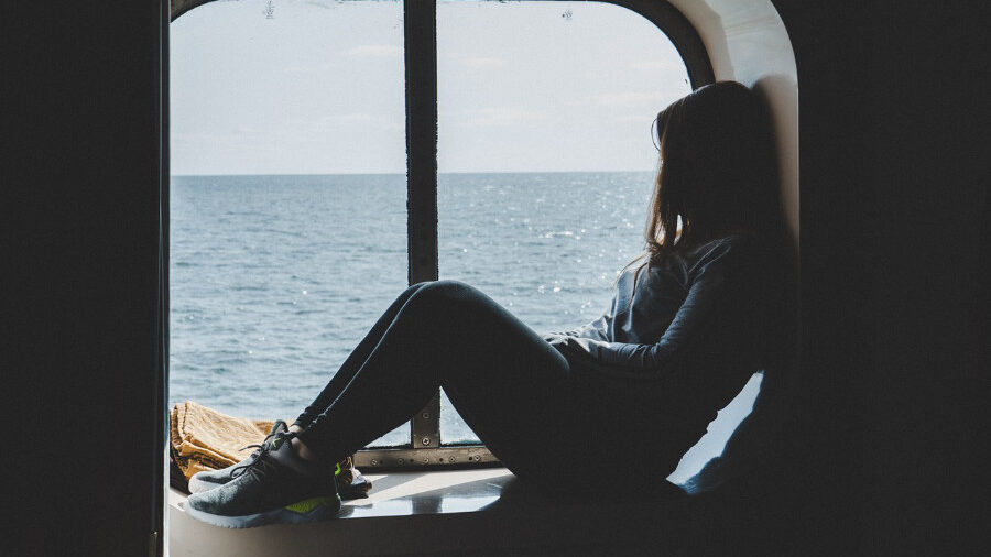 A person sitting by a large window, gazing out at the calm ocean under a bright sky.