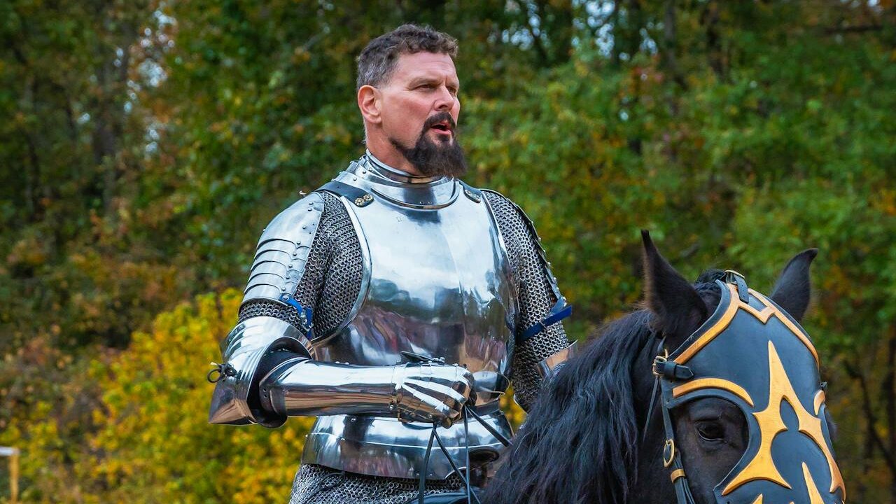 Man dressed in medieval knight armor riding a black horse with decorative headgear.