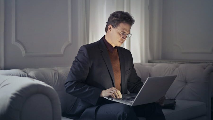 Man in a suit and glasses working on a laptop while seated on a couch in a softly lit room.