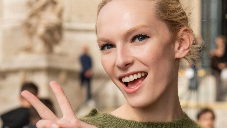 Alex Consani smiling and flashing a peace sign while wearing a striped green sweater at Paris Fashion Week 2024.