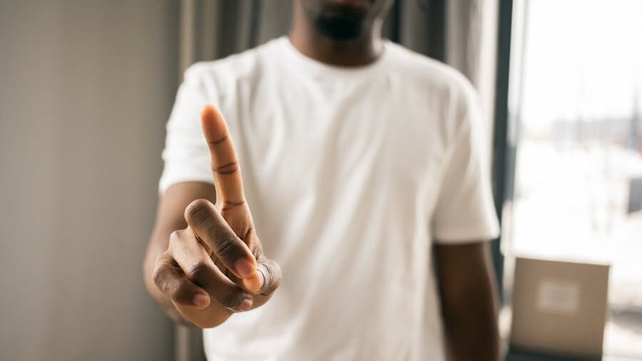 A person in a white t-shirt extending their index finger towards the camera in a direct and assertive gesture, with a blurred indoor background.