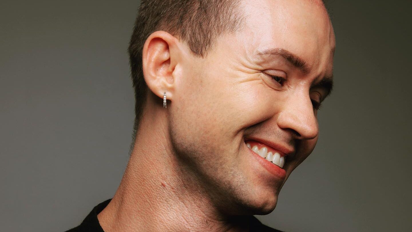 Chase Johnsey tilting his head and smiling, wearing a black outfit in a professional studio setting.