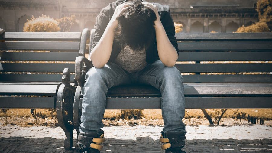 A person sitting on a bench outdoors, leaning forward with their head in their hands, expressing distress or contemplation.