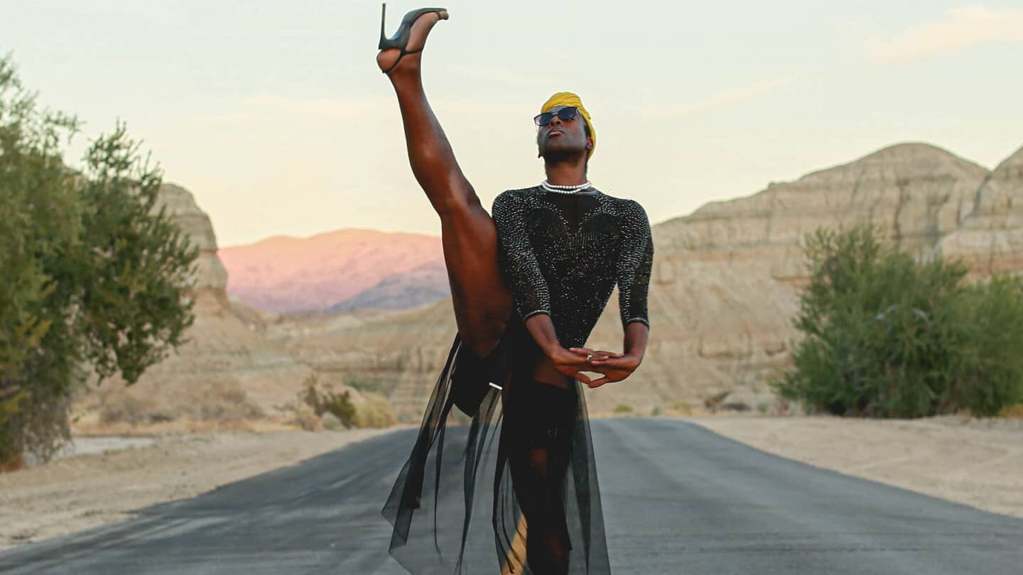 Darling Squire performing a high leg extension on a road at Joshua Tree National Park, wearing a black outfit and heels.