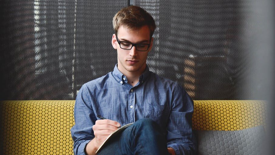 Man wearing glasses and a blue shirt, sitting on a yellow patterned couch, writing in a notebook.