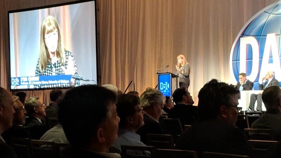 Lynn Ann Conway speaking at the podium during a Plenary Session at D60, with her image projected on a large screen and audience members in the foreground.