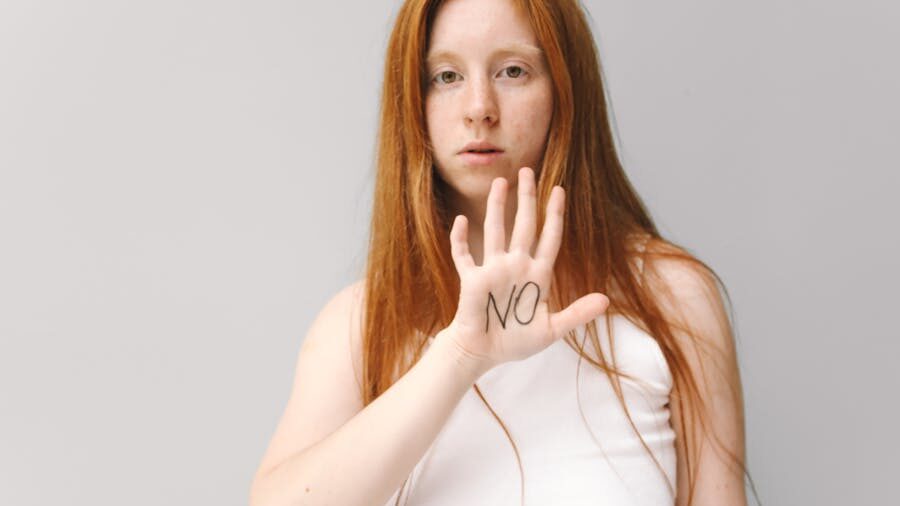 Woman with long red hair wearing a white top, holding up her hand with "NO" written on her palm, against a neutral background.