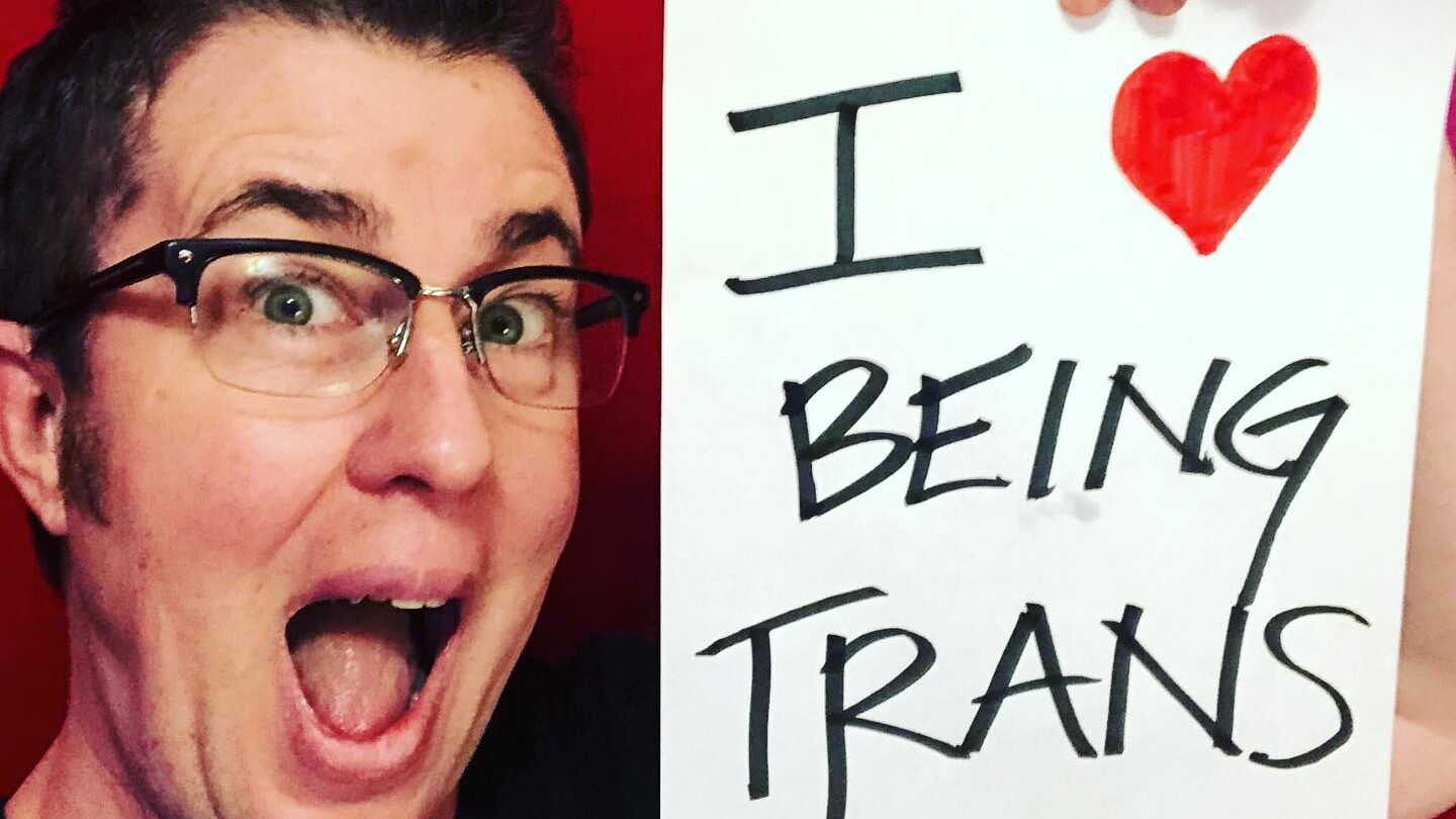 Sean Dorsey smiling and holding a sign that reads "I ❤️ BEING TRANS" against a red background.