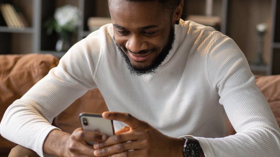 Smiling man wearing a white sweater, sitting on a couch while looking at his smartphone.