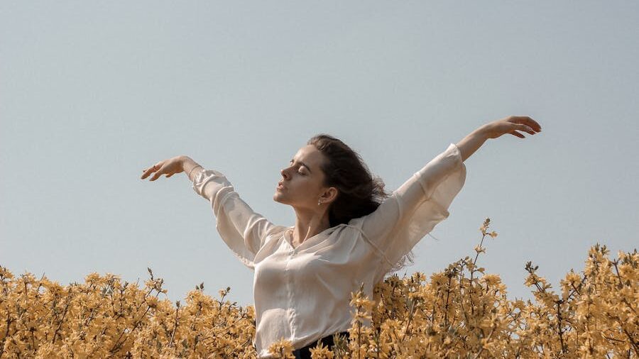 A young woman in a white blouse stands in a field of yellow flowers, arms outstretched, enjoying the warmth of the sun with a peaceful expression.