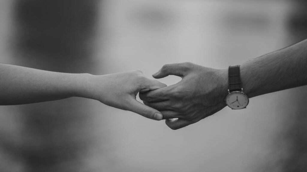 A hand wearing a watch holding another hand in an intimate gesture, in monochrome.