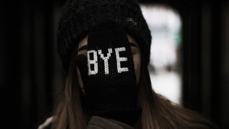 Person wearing a black beanie and gloves covers their face with a gloved hand that has 'BYE' stitched in white letters