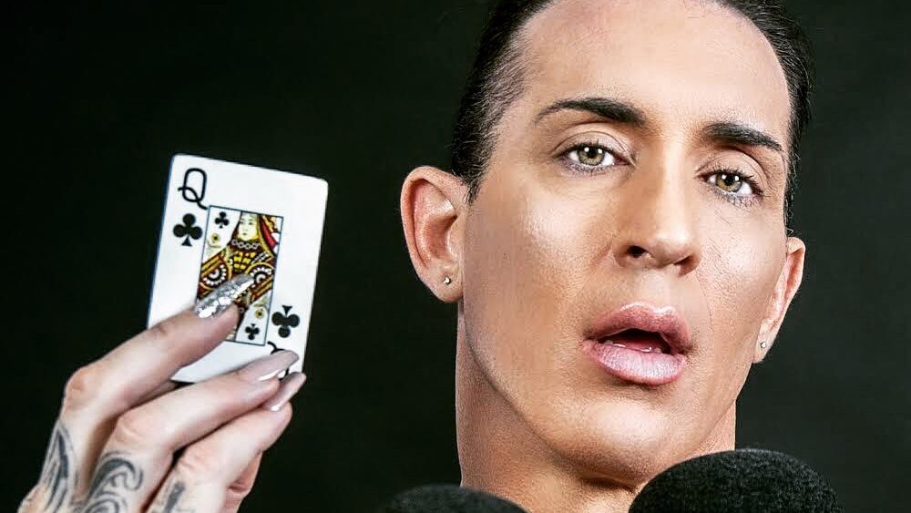 Gabbi Tuft holding a Queen of Clubs playing card, looking into the camera with a bold expression, nails painted and tattooed hand visible.