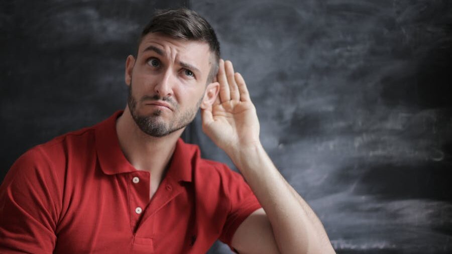 A man in a red polo shirt sitting in front of a blackboard, raising his hand to his ear with a curious expression.