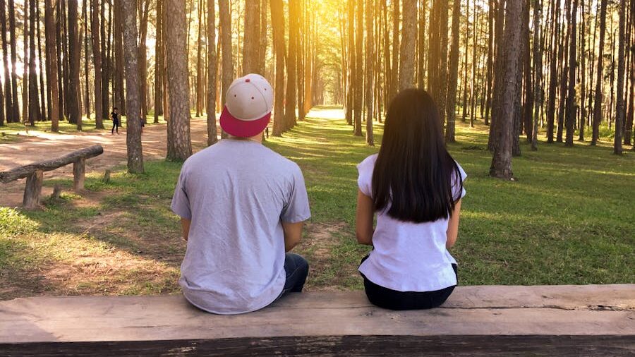 A young man and woman sitting on a wooden bench, facing a sunlit forest path.