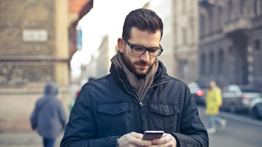 Man with glasses and a beard wearing a dark jacket and scarf, looking at his smartphone on a city street.