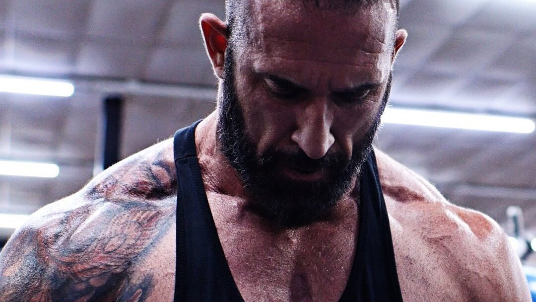 Close-up of Tyler Reks in a gym, wearing a black tank top, looking down with a determined expression, muscles and tattoos visible.