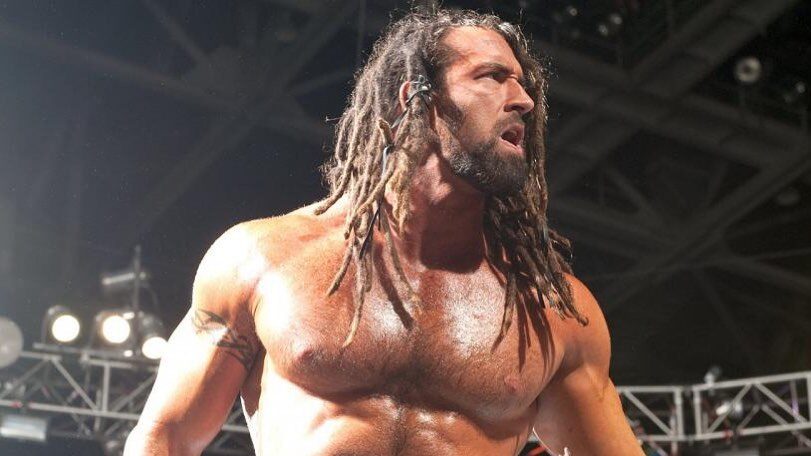 Tyler Reks in a WWE match, shirtless with long dreadlocks, standing under bright arena lights with an intense expression.