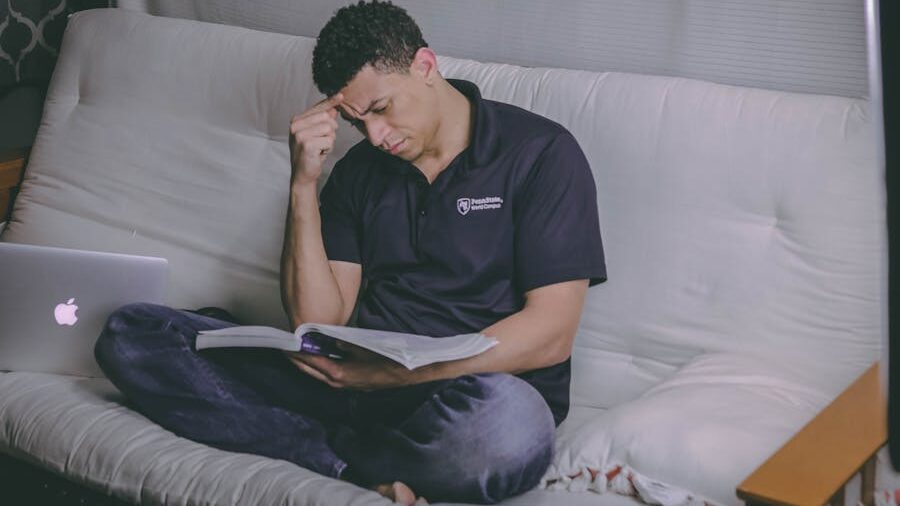 A man sitting on a couch, reading a book with a concentrated expression while holding his forehead. A laptop is placed beside him.