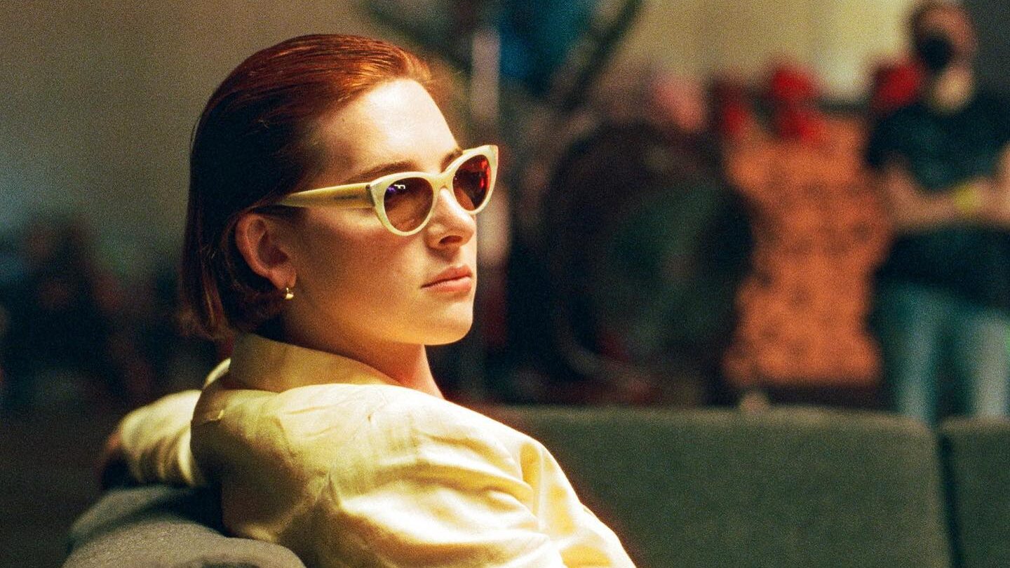 Hari Nef lounging on a gray couch, wearing vintage Gaultier sunglasses and a yellow blazer. The dimly lit background suggests a film set.