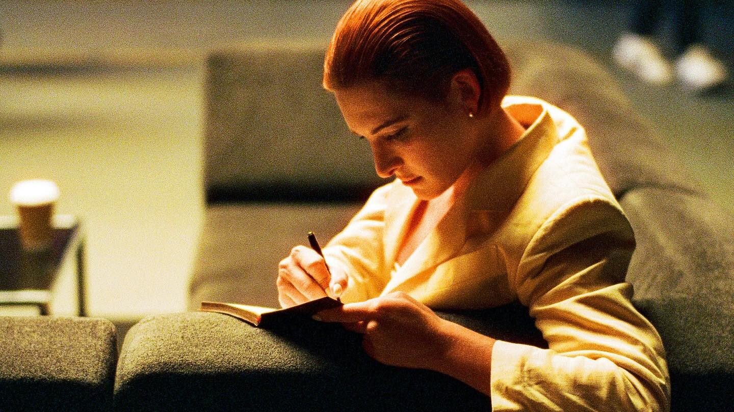 Hari Nef sitting on a gray couch, dressed in a yellow blazer, writing in a notebook on a dimly lit film set.