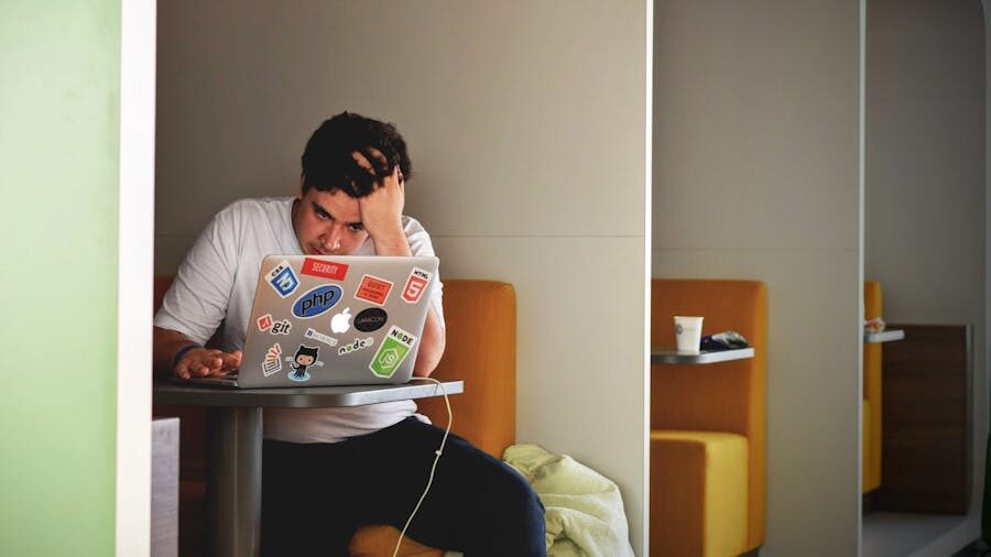 A man sitting in a booth, working on a laptop covered in tech-related stickers, with a stressed expression and his hand on his head.