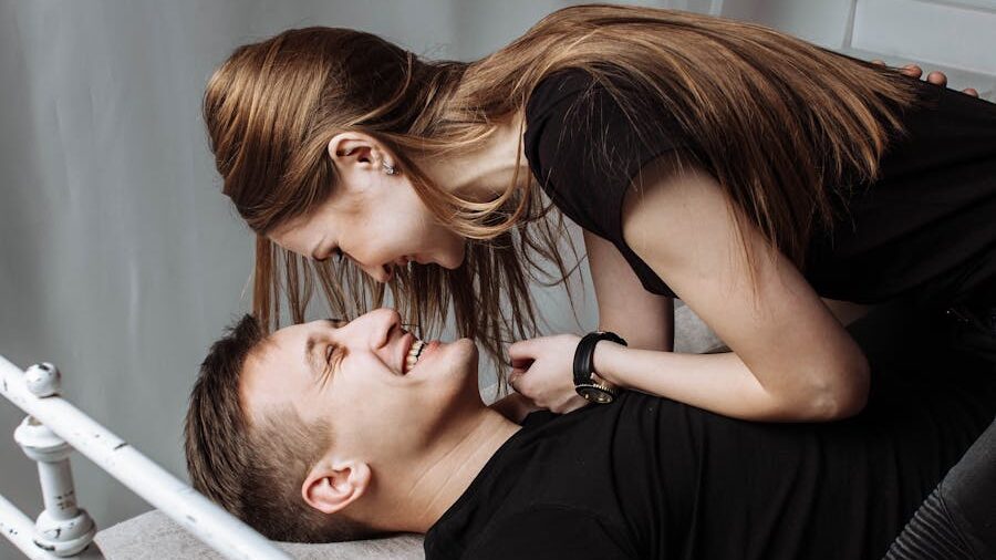 A smiling couple dressed in black, playfully leaning towards each other on a bed.