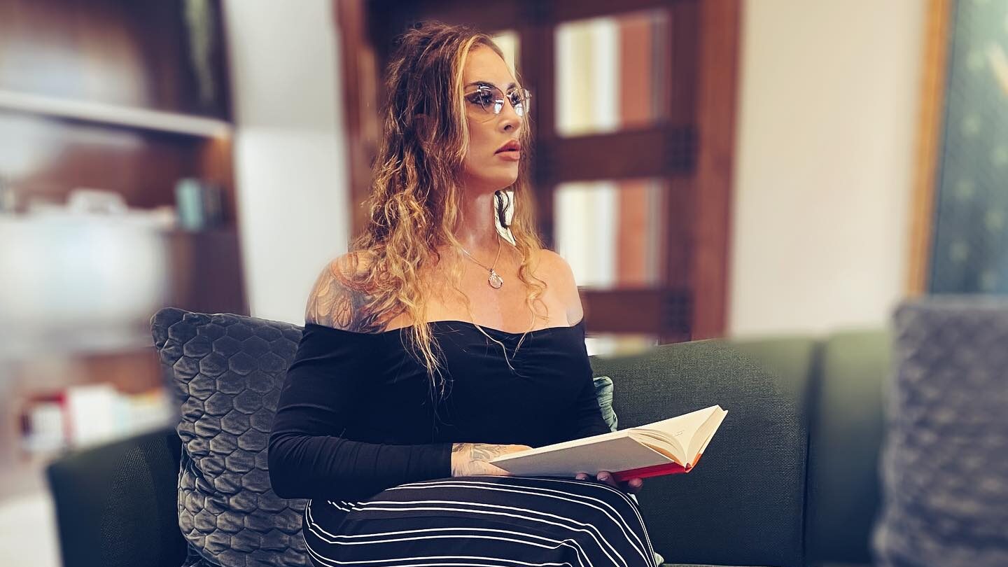 Gabbi Tuft sitting on a green couch in a luxury hotel, wearing glasses and a black off-the-shoulder top, holding an open book.