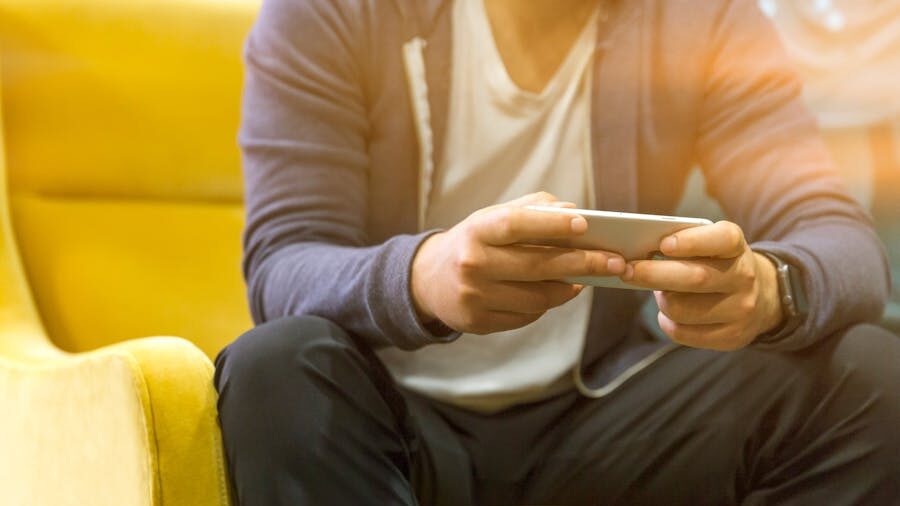 Person in a hoodie and white shirt sitting on a couch, focused on their smartphone.