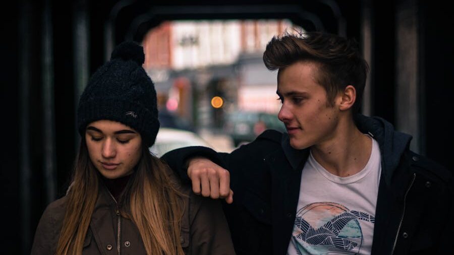 Young man leaning on a young woman’s shoulder as they walk through a dimly lit urban tunnel.