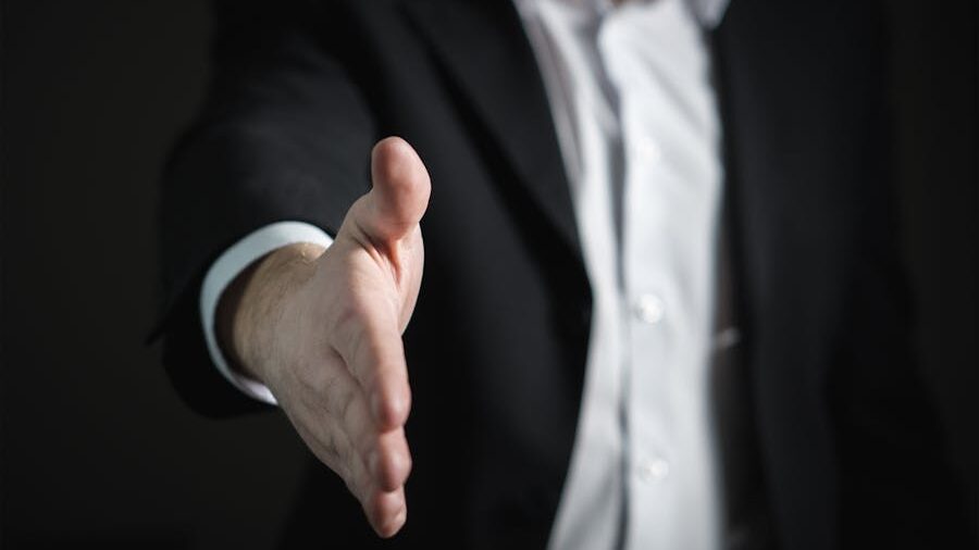 A man in a suit extending his hand for a handshake against a dark background.