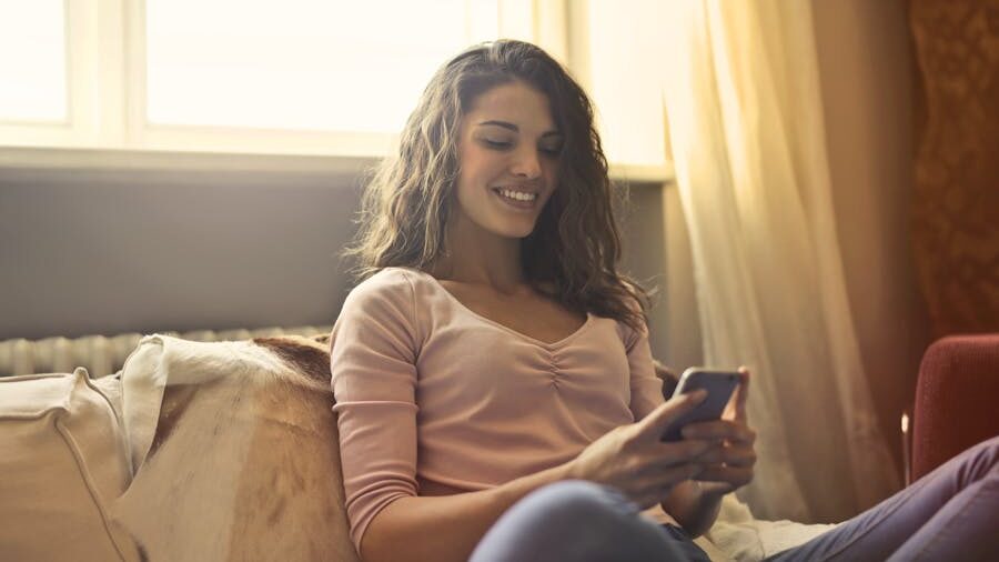  Smiling woman sitting on a couch, looking at her phone in a warmly lit room.