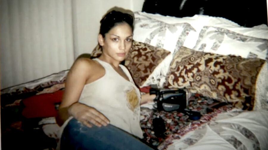 Miriam Rivera lying on a patterned bed, holding a camcorder in a scene from “Miriam: Death of a Reality Star.”