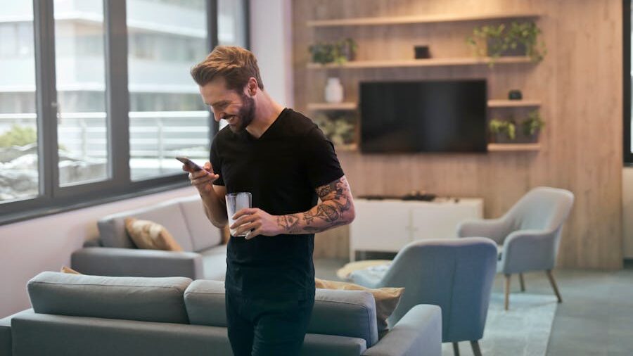 A smiling man in a black shirt looking at his phone while holding a drink, walking through a modern, cozy apartment.