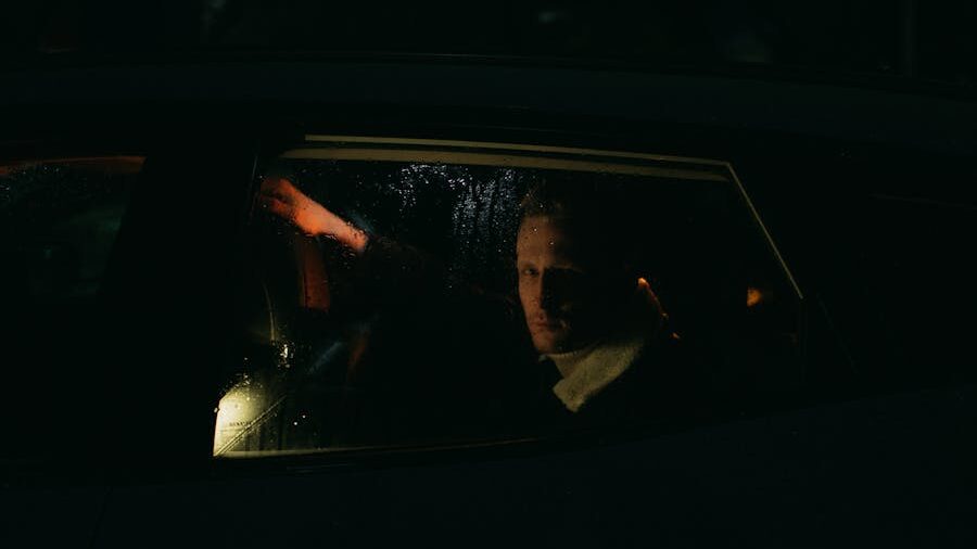 A man sits in the backseat of a car at night, his face dimly lit as he looks out the rain-covered window with a serious expression.