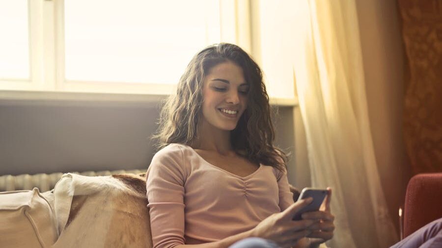 Smiling woman sitting on a sofa, wearing a light pink top, looking at her phone in soft natural light from a window.