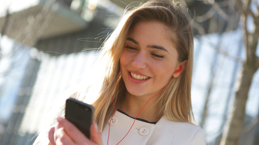 Blonde woman smiling while listening to something on her phone with red earphones outdoors.